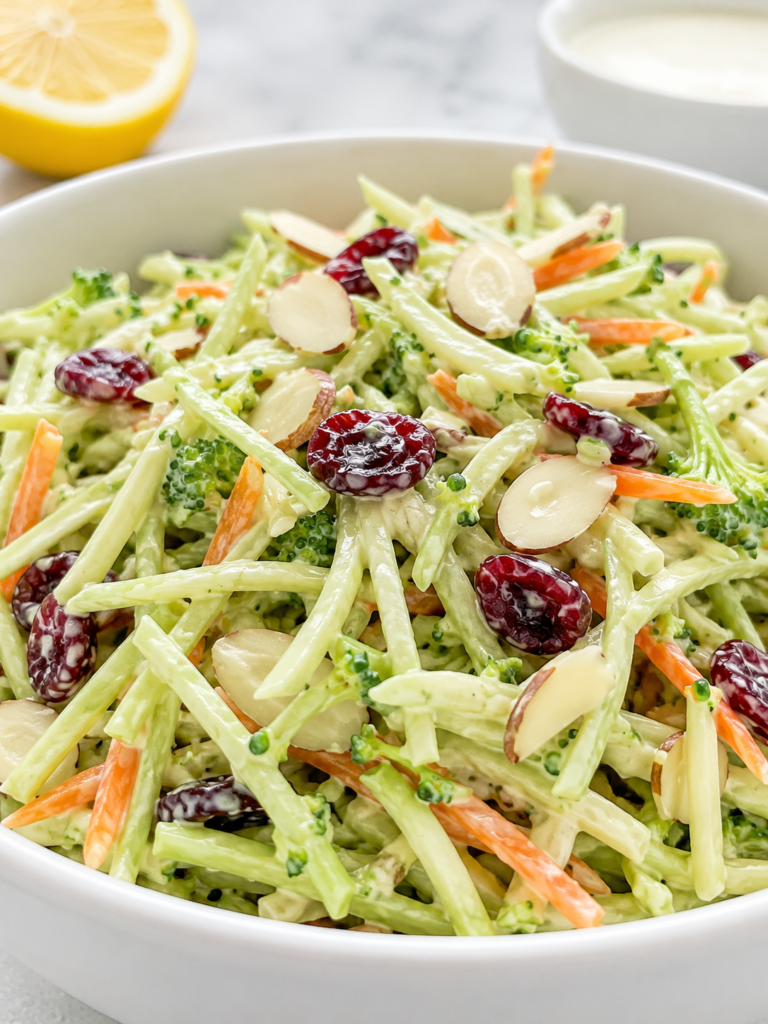 Close-up of creamy broccoli slaw with shredded broccoli stems, carrots, dried cranberries, and sliced almonds in a white bowl.