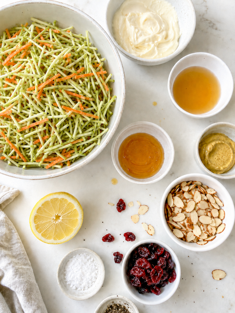 Ingredients for broccoli slaw recipe including broccoli slaw mix, mayo, apple cider vinegar, honey, Dijon mustard, lemon, almonds, and dried cranberries.