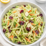 Overhead view of creamy broccoli slaw in a white bowl with dried cranberries, sliced almonds, lemon, and dressing nearby.