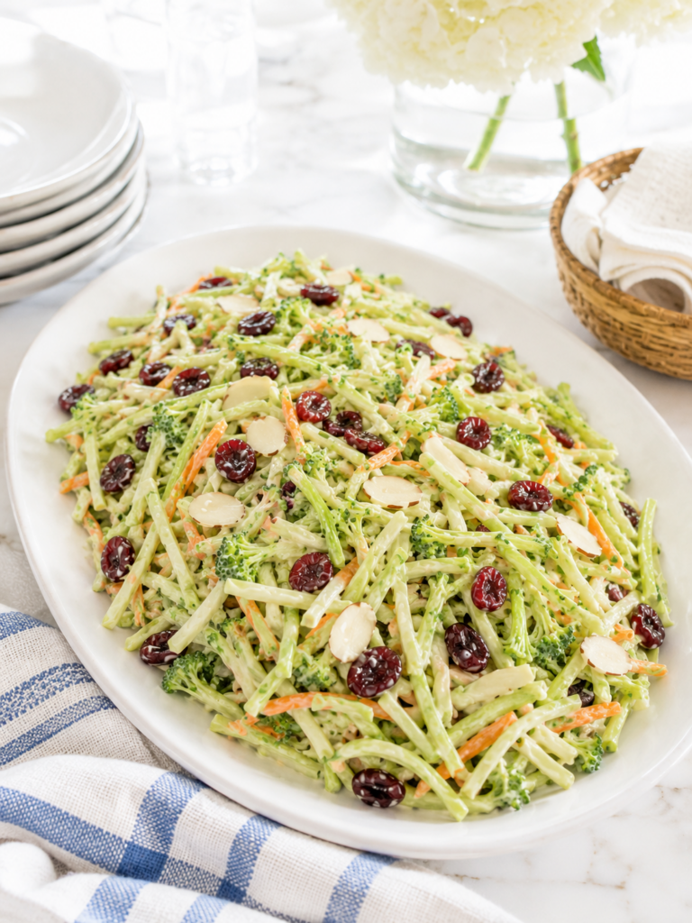 Creamy broccoli slaw served on a white platter at a summer BBQ table with sliced almonds and dried cranberries.