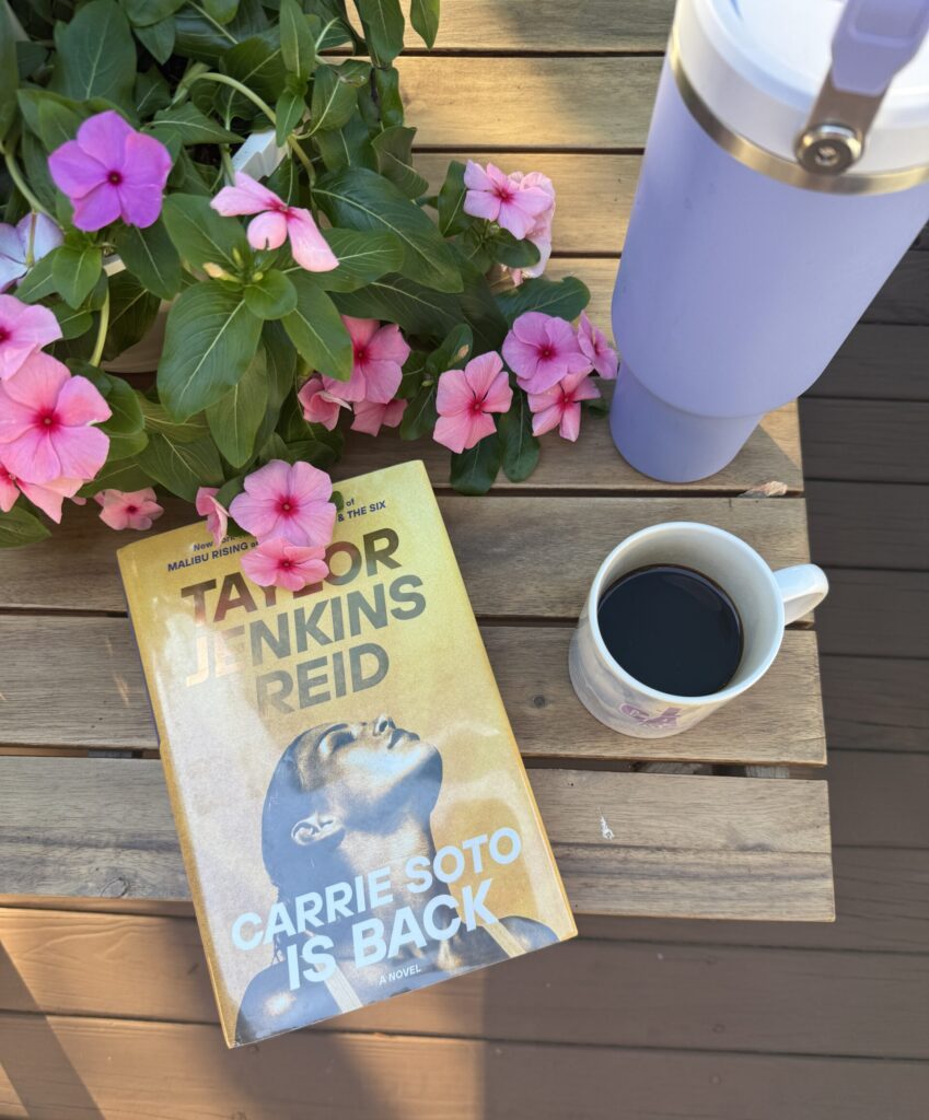A cozy, outdoor reading setup featuring 'Carrie Soto Is Back' by Taylor Jenkins Reid, accompanied by a cup of coffee, purple water bottle, and blooming pink and purple pansy flowers.