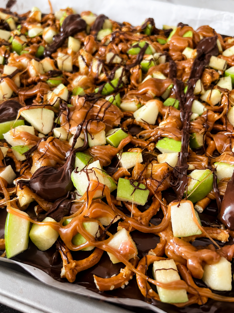 Close-up of caramel apple bark layered with chopped green apples, pretzels, chocolate, and caramel drizzle.