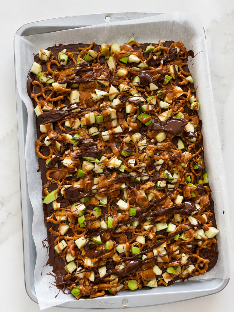 Overhead view of homemade caramel apple bark with pretzels and green apples on a parchment-lined baking sheet.