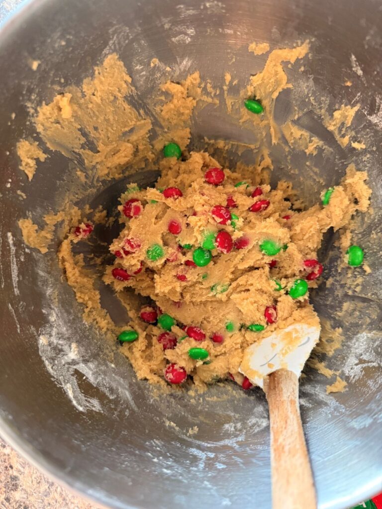 Using a spatula to fold in red and green m&m candies into cookie dough in kitchen aid mixing bowl
