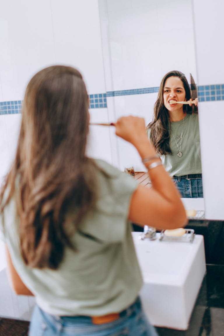 A young woman in casual attire brushing her teeth while looking in the bathroom mirror.