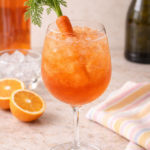 glass of aperol spritz garnished with a carrot fand ingredients in the background for a fun twist on an easter cocktail.