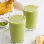 two glasses of matcha smoothie with banana and match powder on marble countertop