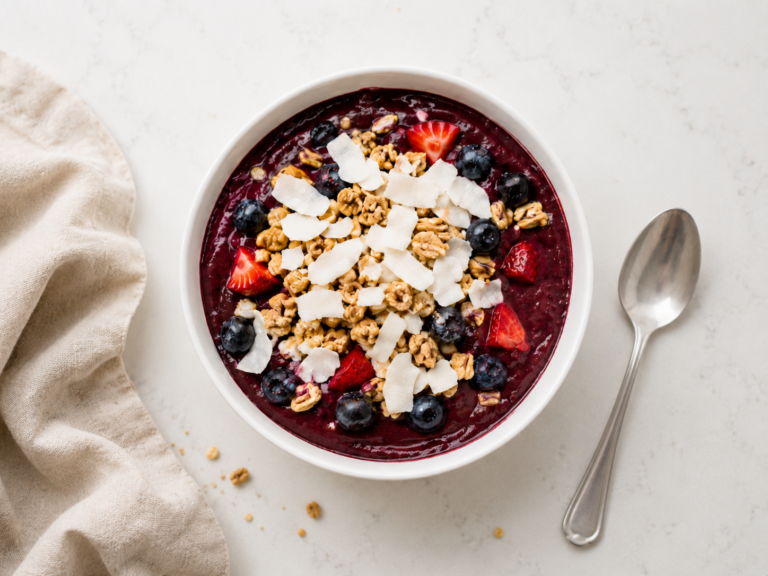Overhead view of an acai bowl in a white ceramic bowl topped with granola, coconut flakes, blueberries, and strawberries on a light countertop.