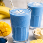Bright blue spirulina smoothies in clear glasses with tropical fruit on a light marble countertop