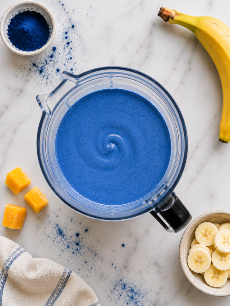 Blue spirulina smoothie blended in a clear blender on a marble countertop with blue spirulina powder and tropical fruit nearby