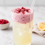 glass of refreshing lemonade topped with cherry cold foam on marble countertop with lemons and small bowl of dried cherries