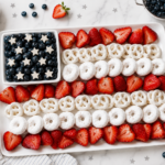 Easy 4th of July fruit plate arranged on a white rectangular serving tray with strawberries, blueberries, yogurt-covered pretzels, and powdered donuts styled like an American flag.