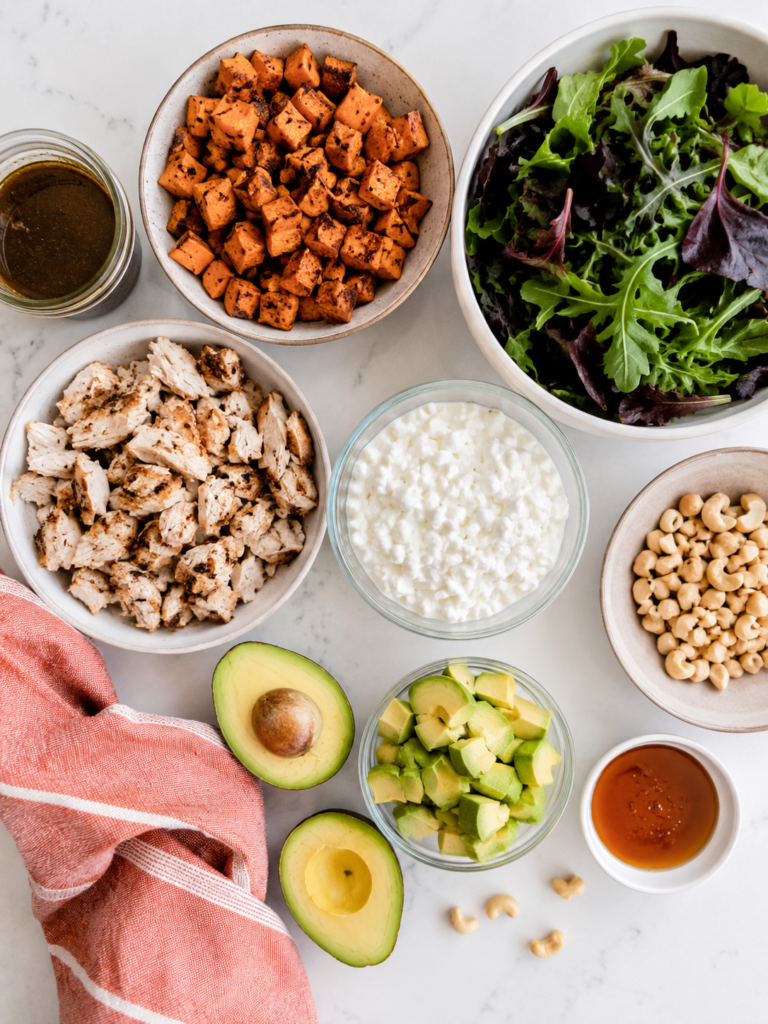 Ingredients for a hot honey cottage cheese bowl including roasted chicken, sweet potatoes, mixed greens, avocado, cottage cheese, cashews, balsamic vinaigrette, and hot honey.