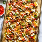 Loaded zucchini nachos with taco meat, melted cheese, diced tomatoes, jalapeños, and creamy drizzle on a parchment-lined sheet pan.