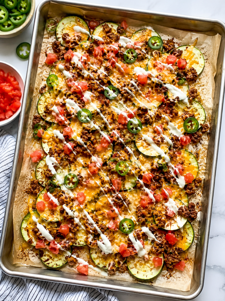 Loaded zucchini nachos with taco meat, melted cheese, diced tomatoes, jalapeños, and creamy drizzle on a parchment-lined sheet pan.