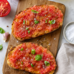 Overhead view of pan con tomate made with grated tomatoes on toasted sourdough bread with olive oil, flaky sea salt, black pepper, and fresh basil on a wooden cutting board.