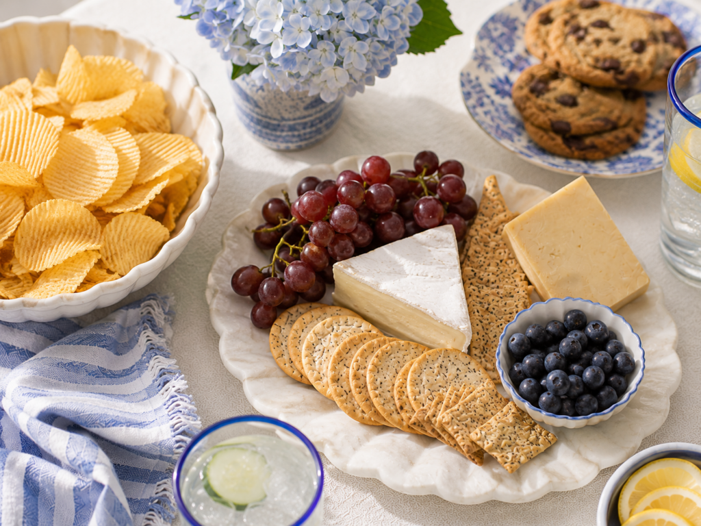 Cheese, crackers, grapes, blueberries, chips, and chocolate chip cookies styled on pretty serving dishes with blue accents for an easy summer snack setup.