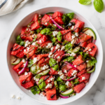 Overhead view of watermelon feta salad with basil in a white serving bowl, topped with cucumber, red onion, feta, and balsamic drizzle.
