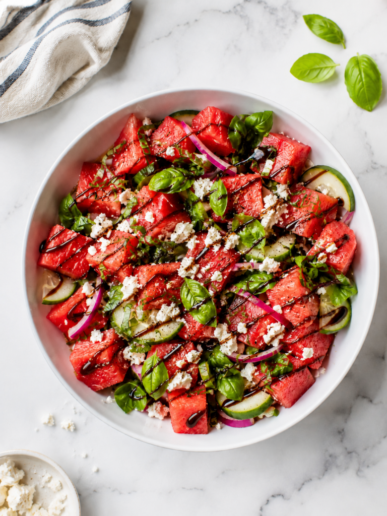 Overhead view of watermelon feta salad with basil in a white serving bowl, topped with cucumber, red onion, feta, and balsamic drizzle.