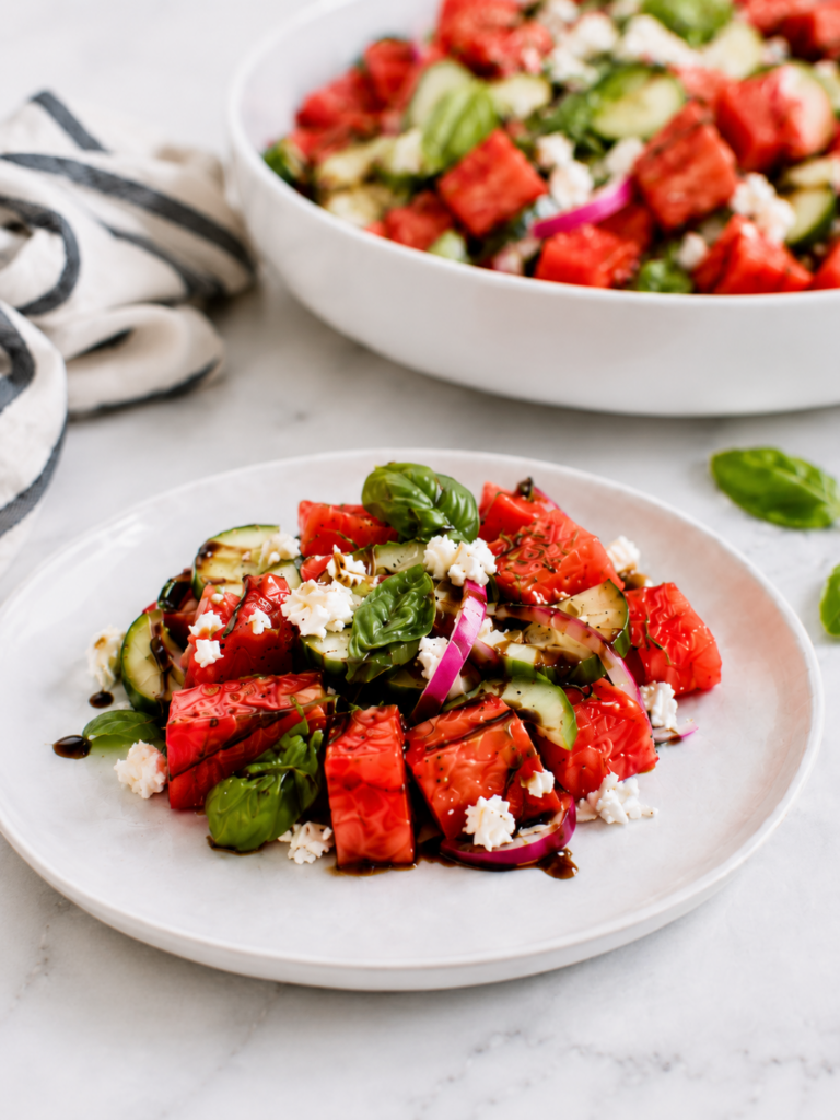 Plated watermelon feta salad with basil topped with cucumber, feta, red onion, and balsamic drizzle, with the serving bowl softly blurred in the background.