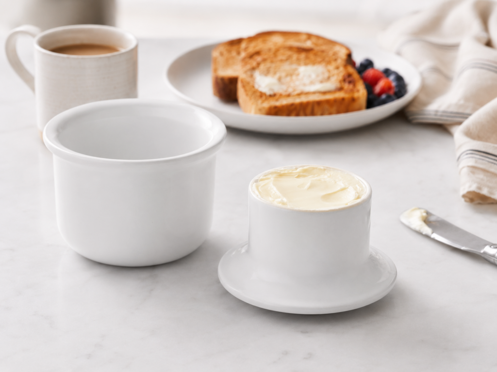 White ceramic butter keeper filled with soft butter on a marble countertop with toast, coffee, and breakfast items blurred in the background.