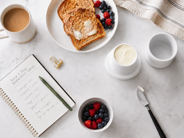 Overhead view of a white ceramic butter keeper with soft butter, toast, berries, coffee, and a butter knife on a white marble countertop.