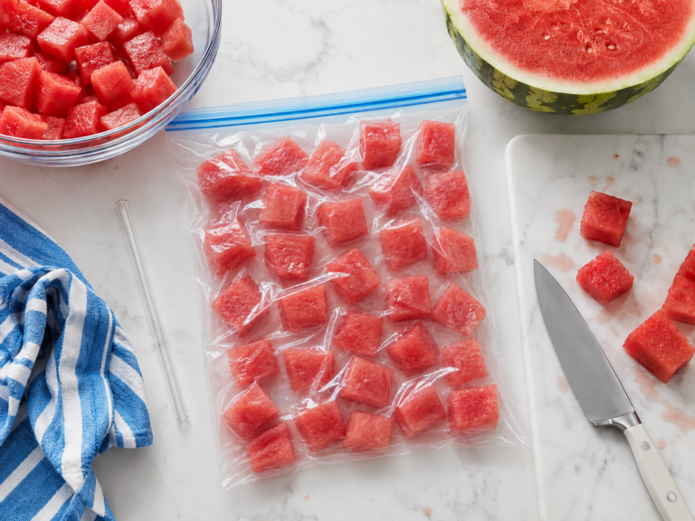 Overhead view of fresh watermelon being prepped for freezing on a white marble countertop. A clear zip-top freezer bag filled with juicy watermelon cubes arranged in a single layer sits in the center, with the fruit visibly sealed inside the bag. Surrounding it are a glass bowl of extra watermelon cubes, a halved watermelon, a marble cutting board with a kitchen knife and a few cut pieces, subtle watermelon juice drips, a clear straw, and a blue-and-white striped kitchen towel, creating a bright, practical summer kitchen scene.