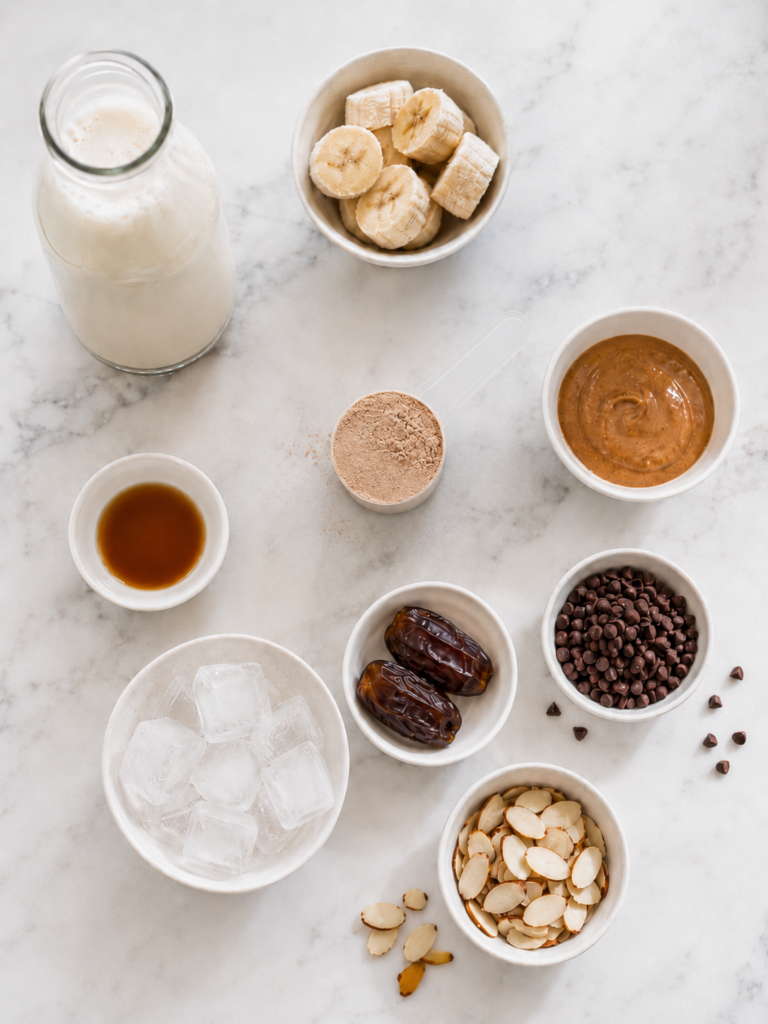 Ingredients for a chocolate protein smoothie including almond milk, frozen banana, protein powder, almond butter, dates, ice, chocolate chips, and sliced almonds.