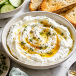 Homemade Greek yogurt cream cheese topped with olive oil, honey, herbs, flaky salt, and black pepper, served with toasted sourdough and cucumber slices.