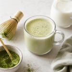 Healthy matcha green tea latte in a clear glass mug with matcha powder, bamboo whisk, milk, and sage towel on a light marble countertop.