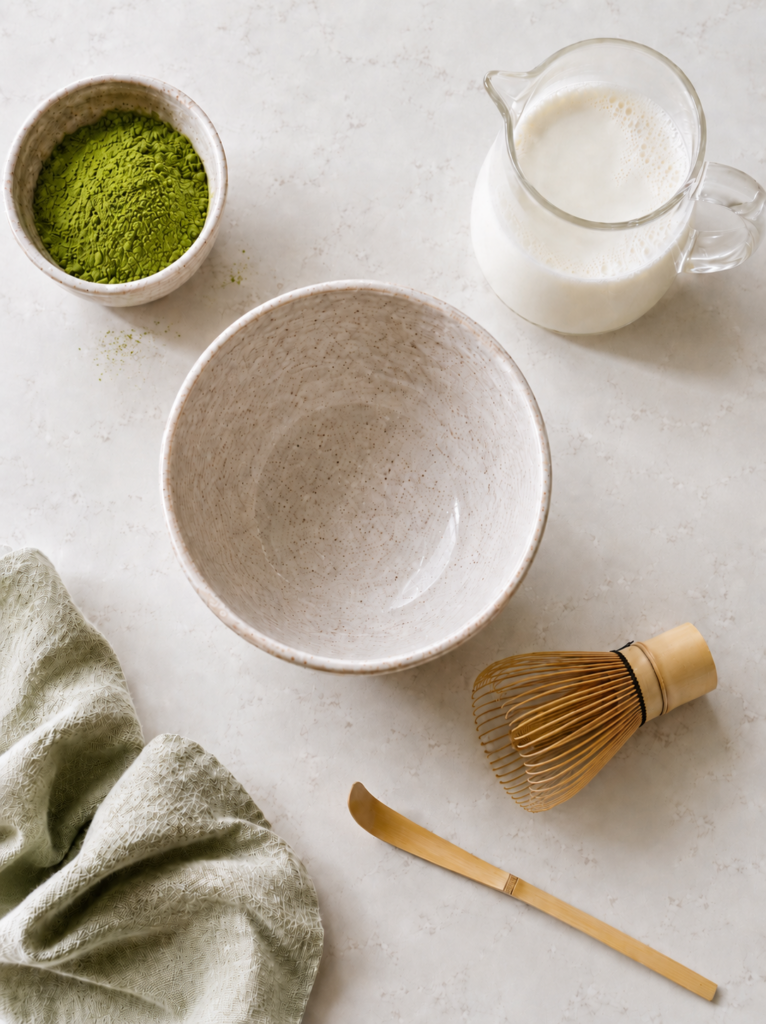 Matcha latte ingredients and tools arranged on a light countertop with matcha powder, bamboo whisk, milk pitcher, ceramic bowl, and sage towel.