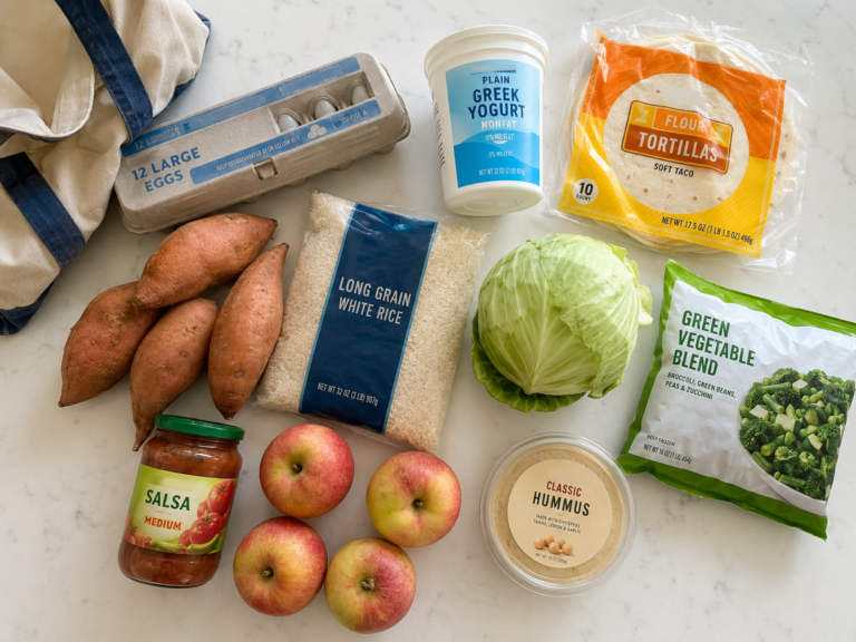 Overhead grocery haul on a white quartz countertop with sweet potatoes, eggs, Greek yogurt, tortillas, rice, cabbage, frozen vegetables, salsa, apples, hummus, and a reusable shopping bag.