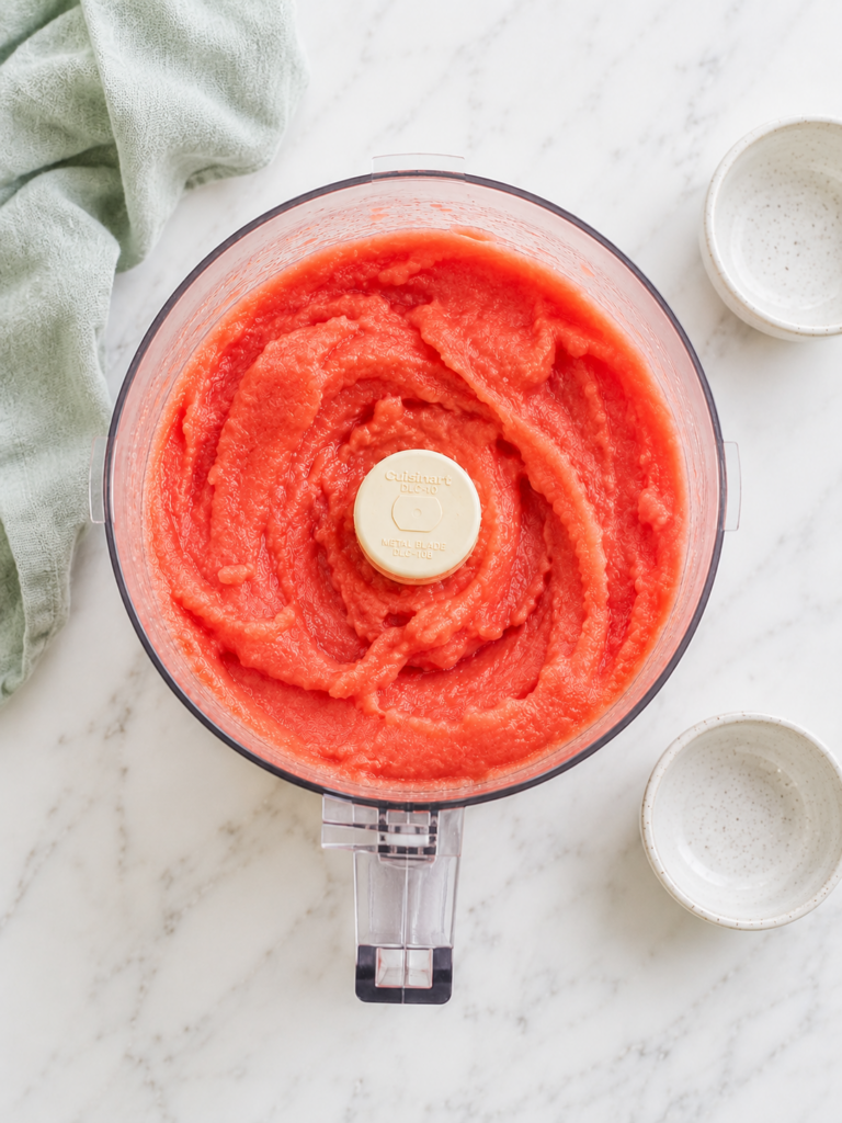 Overhead view of watermelon sorbet blended in a food processor with empty ingredient bowls nearby.