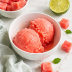 Watermelon sorbet scooped in a white bowl with frozen watermelon cubes and lime on a marble countertop.