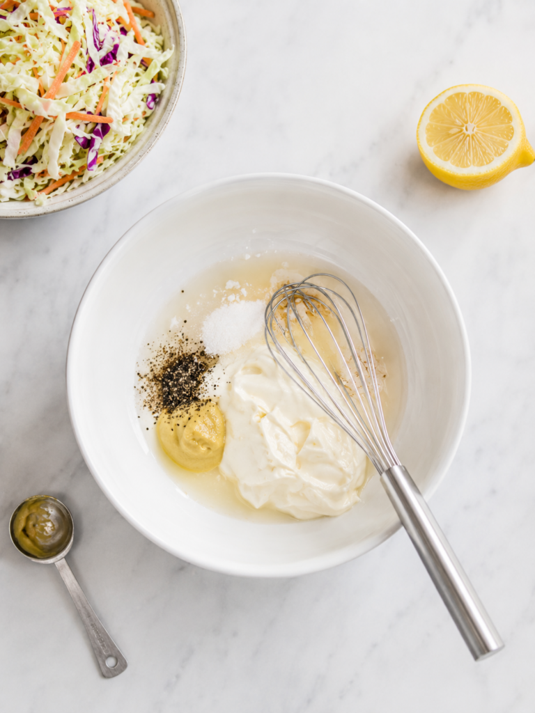 Coleslaw dressing ingredients added to a white bowl before whisking, with bagged slaw mix nearby.