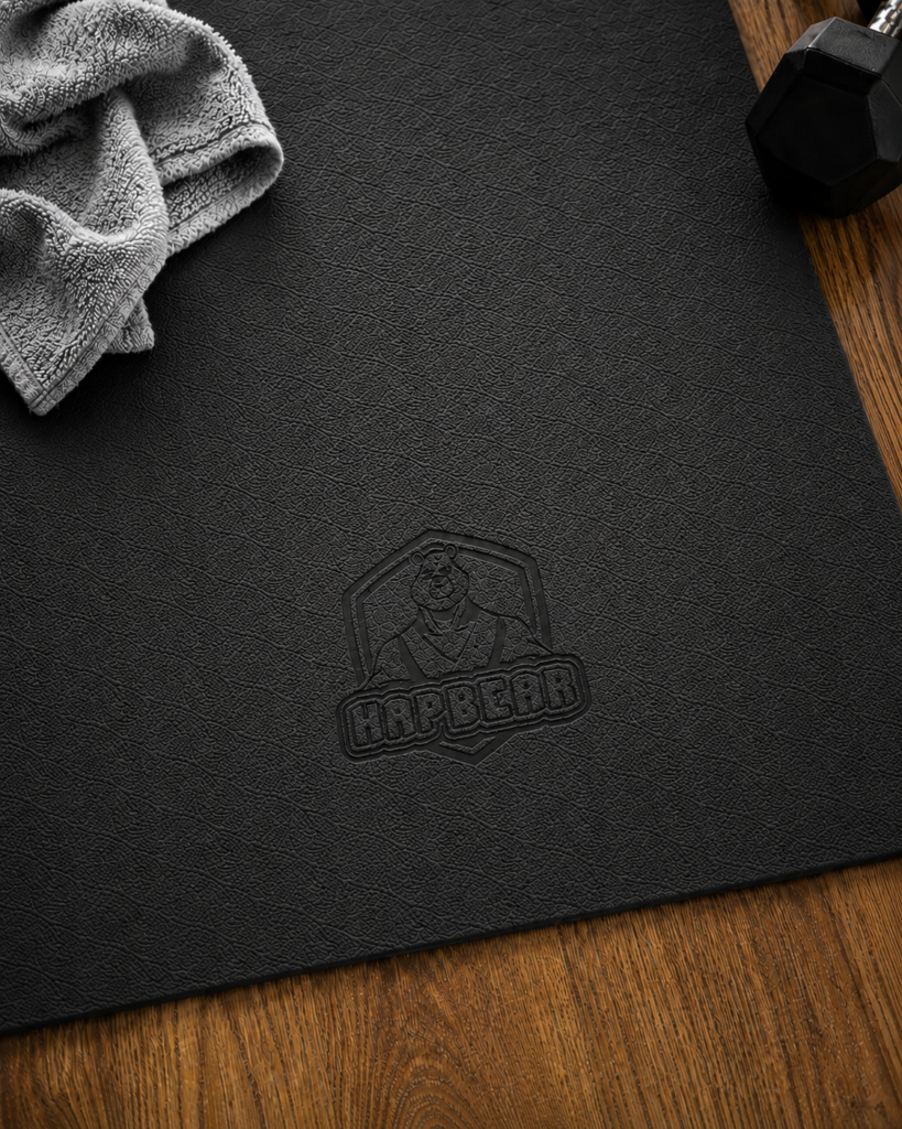 Close-up of the black Hapbear extra large yoga mat showing the textured surface, embossed logo, a small gray hand towel, and a dumbbell on wood flooring.