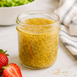 Homemade Panera poppyseed dressing in a glass jar with visible poppy seeds, beside fresh strawberries and a bowl of lettuce