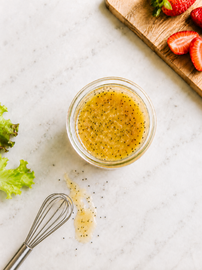 Copycat Panera poppyseed dressing in a jar, shot from above with a whisk, fresh strawberries, and green lettuce on a marble counter