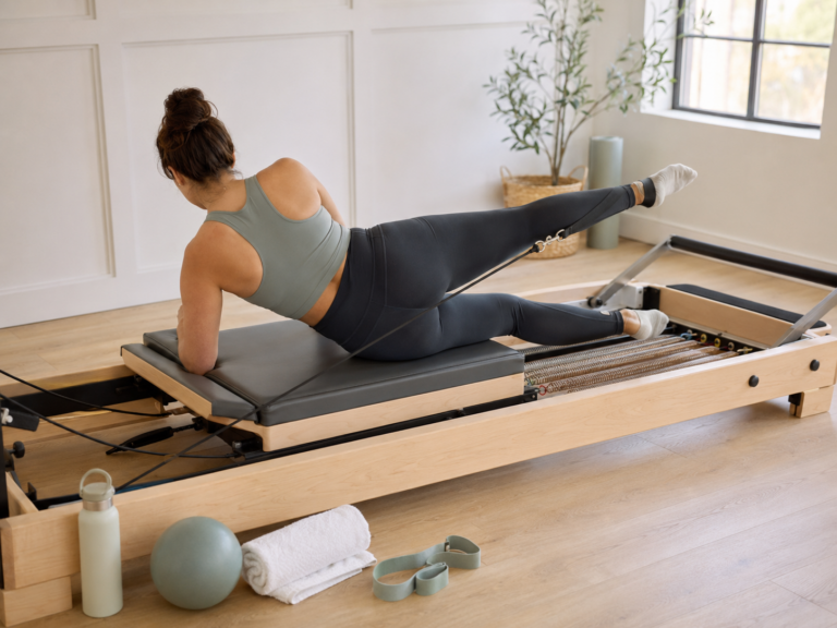 Woman doing a side-lying leg exercise on a reformer Pilates machine in a bright studio with a towel, water bottle, resistance band, and small Pilates ball nearby.