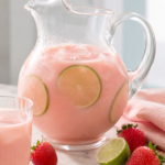Creamy pink strawberry Brazilian lemonade with lime slices and ice in a clear glass pitcher on a marble countertop.