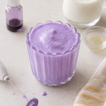 Homemade ube cold foam in a tall ribbed flower glass with a dirty frother, ube extract, cream, vanilla syrup, and striped towel on a light countertop.