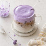 Iced latte topped with thick purple ube cold foam in a clear glass mug with a glass straw, styled on a marble coaster with a frother, small ribbed glass of leftover foam, and a striped towel on a light countertop.