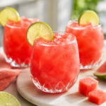 Three short glasses of watermelon agua fresca with ice and lime wheels served on a marble tray with fresh watermelon cubes and lime on a white marble countertop.