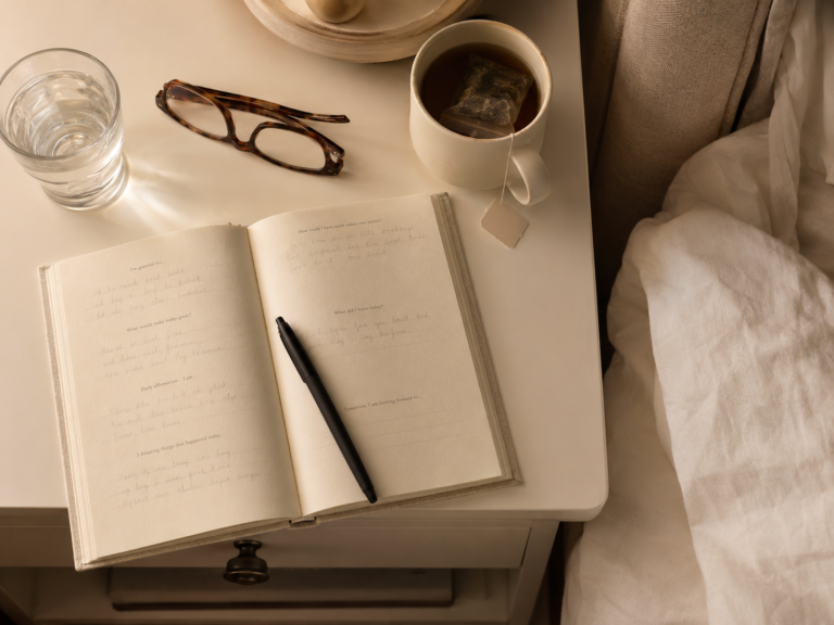 Open guided journal with a black pen, herbal tea, water and reading glasses on a cream nightstand beside a softly rumpled bed.